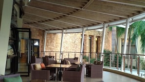 Een overdekt terras met rieten meubels en een glazen balustrade, met uitzicht op een bakstenen gebouw en palmbomen.