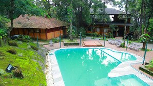 Una piscina de borde ondulado, rodeada de abundante vegetación y un pequeño edificio con techo de tejas.