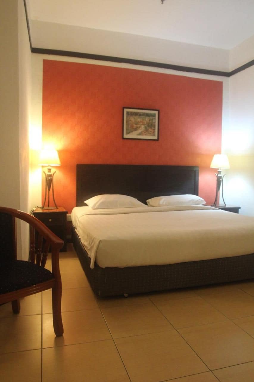 A hotel room with a bed, two bedside lamps, a chair, and a painting on the wall.