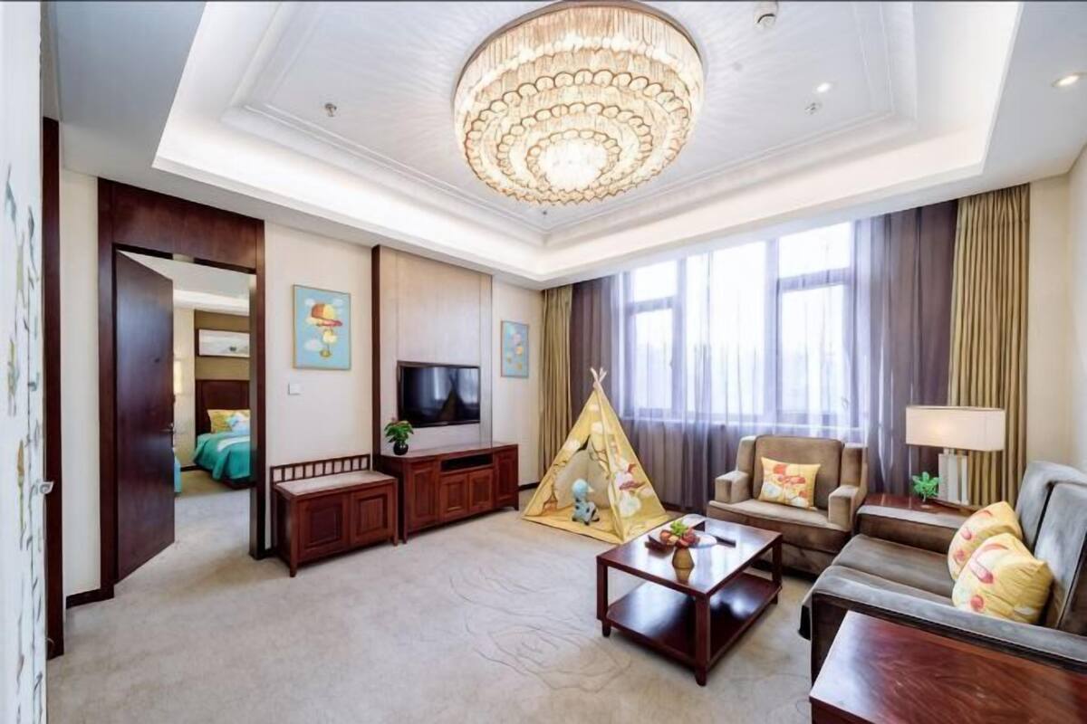 A spacious living room with a chandelier, a television, a sofa, a coffee table, and a window with curtains.