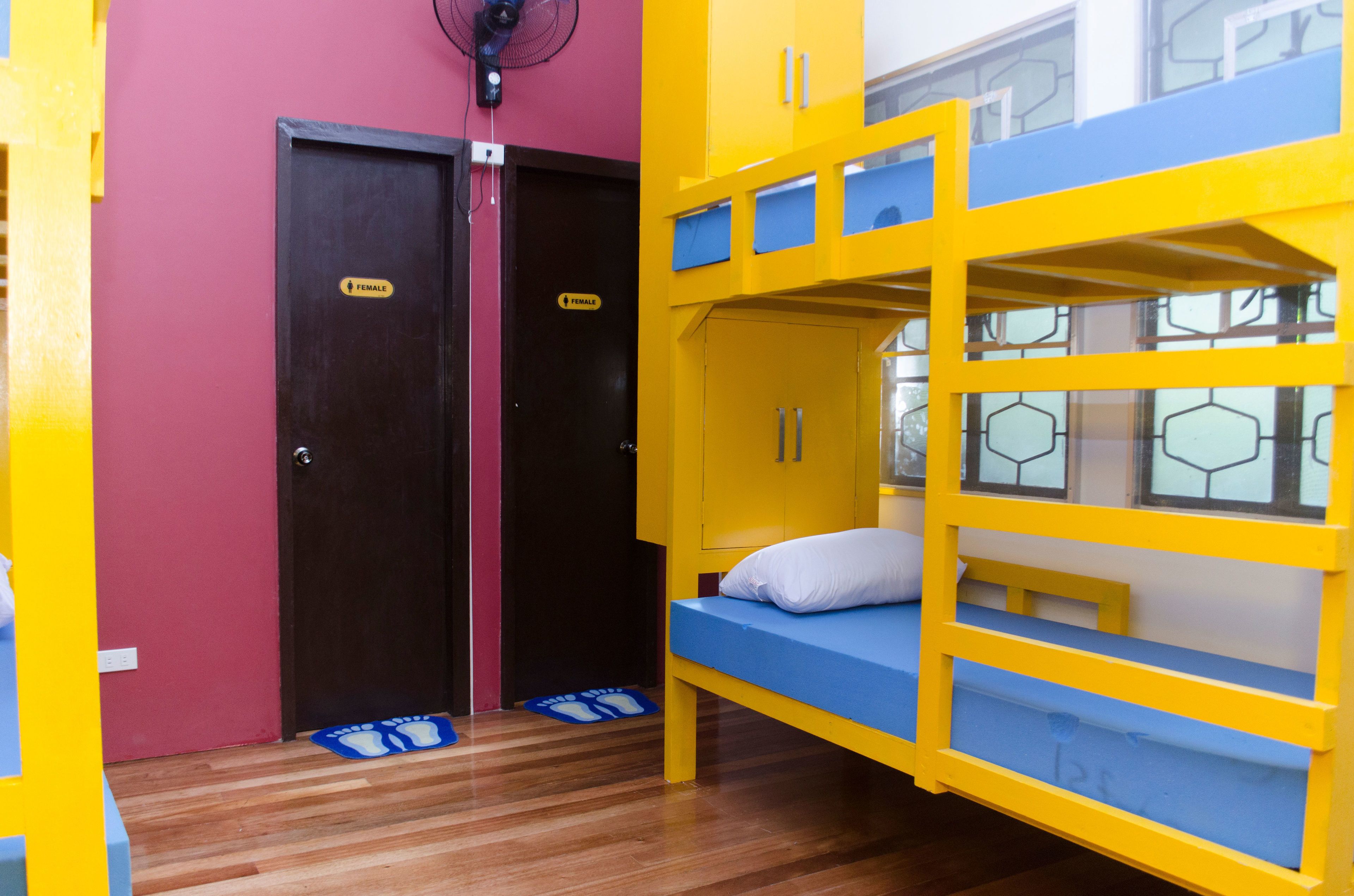 A bunk bed room with a blue mattress and yellow frame, a pair of doors with a 'female' sign, and a fan on the wall.