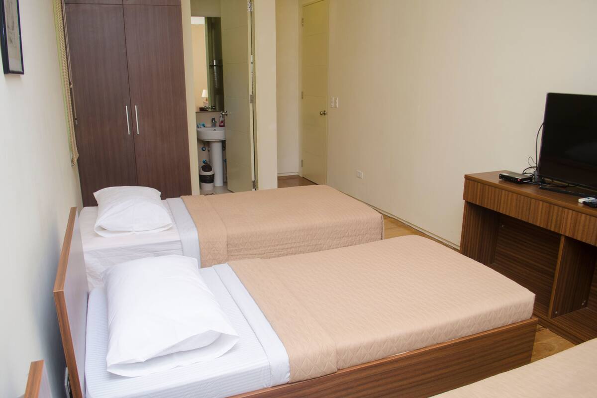 Une chambre avec deux lits simples, un bureau en bois avec un ordinateur, une armoire et une salle de bain visible à travers une porte ouverte.