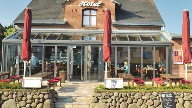 A hotel with a glass facade, outdoor seating, and a stone wall in front. - Hotel Kiose - Landhaus JOJO (Wenningstedt-Braderup)