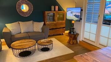 Ein Wohnzimmer mit einer Couch, zwei runden Couchtischen, einer Holzkabine, einem Fernseher und einem Fenster mit Jalousien.