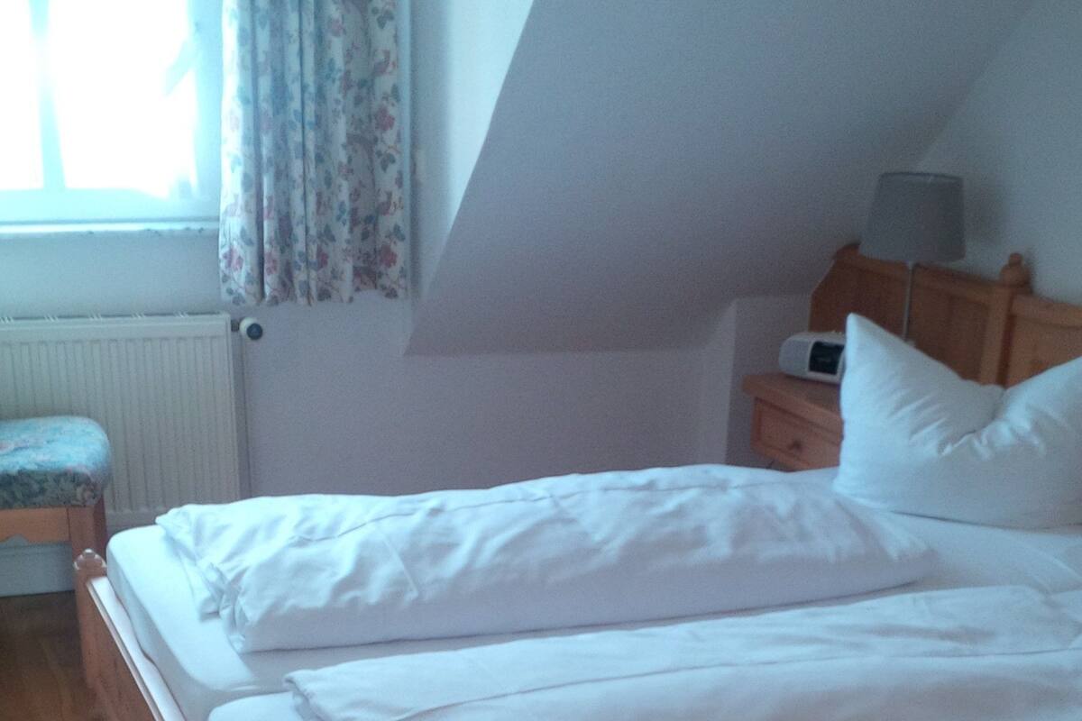 A bedroom with a wooden bed, white bedding, a nightstand with a lamp, and a window with curtains.