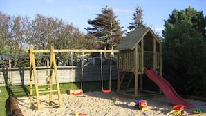 A wooden play structure with a slide and swings, surrounded by sand and greenery. - Hotel Kiose - Landhaus JOJO (Wenningstedt-Braderup)