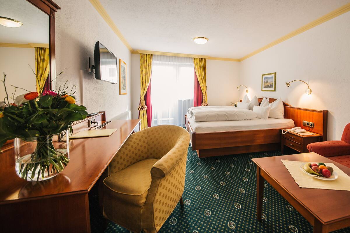 Une chambre d’hôtel avec un lit, un bureau, une chaise, un canapé, une table avec des fruits, un vase avec des fleurs, une télévision et des rideaux.