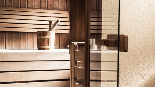 A wooden bench with a wooden bucket and a metal container, likely for holding towels or toiletries, in a room with a wooden wall and a glass door. - Hotel Kristall (Sankt Anton am Arlberg)