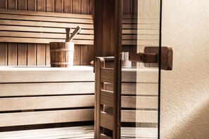 A wooden bench with a wooden bucket and a metal container, likely for holding towels or toiletries, in a room with a wooden wall and a glass door. - Hotel Kristall (Sankt Anton am Arlberg)