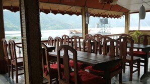 Un restaurante con mesas y sillas de madera, cojines rojos y vistas a un cuerpo de agua y montañas.