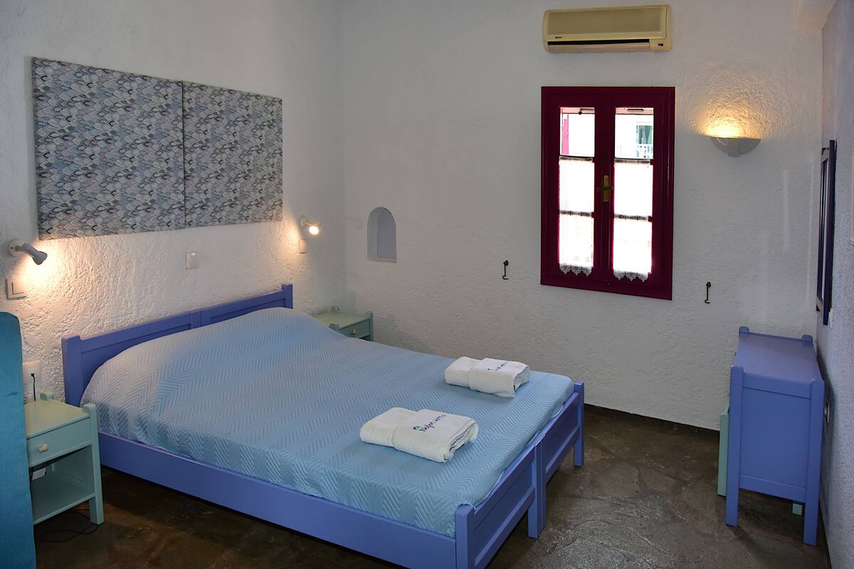 Une chambre avec un lit simple, une table de chevet, un climatiseur fixé au mur, une petite fenêtre avec un cadre rouge et un luminaire fixé au mur.