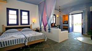 A bedroom with a bed, a stone floor, a window with curtains, a nightstand, and a small table with a lamp. - Hotel Nefeli (Leros)