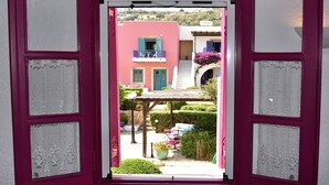 An open window with lace curtains, revealing a courtyard with a table, chairs, and potted plants. - Hotel Nefeli (Leros)