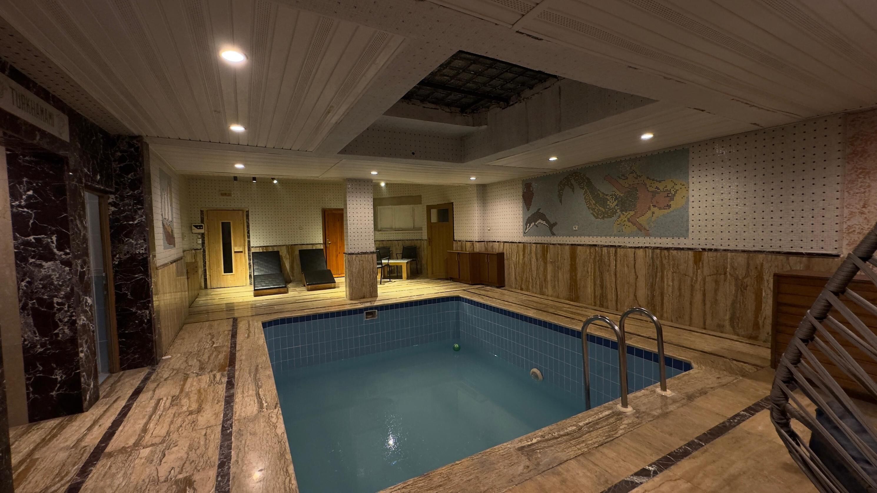 A large indoor swimming pool with a tiled floor, a wooden deck, and a seating area with chairs and a table.