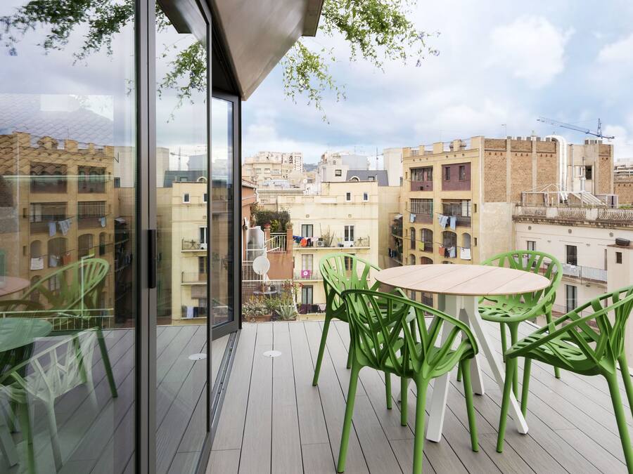 Un balcón con pared de vidrio, sillas verdes y una mesa redonda.