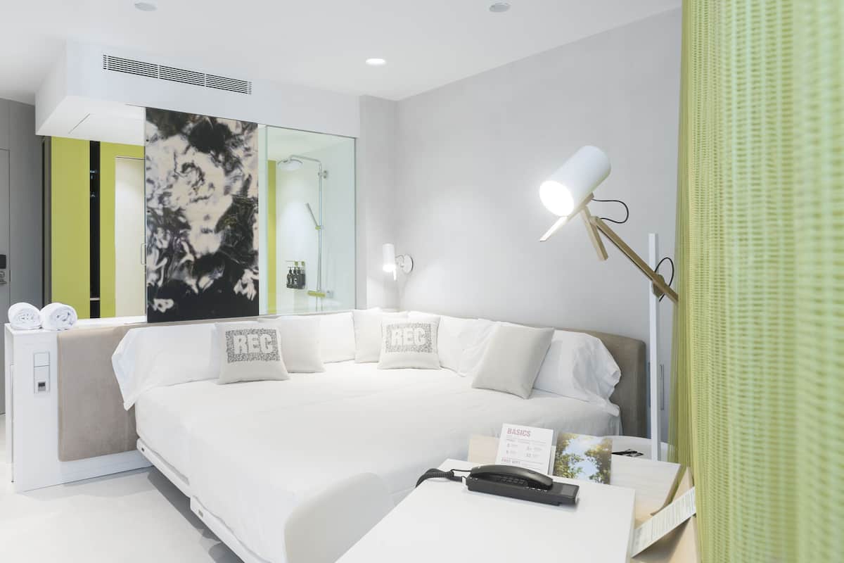 A modern hotel room with a large bed, a bedside table with a phone and a notepad, a wall-mounted mirror, and a green curtain.