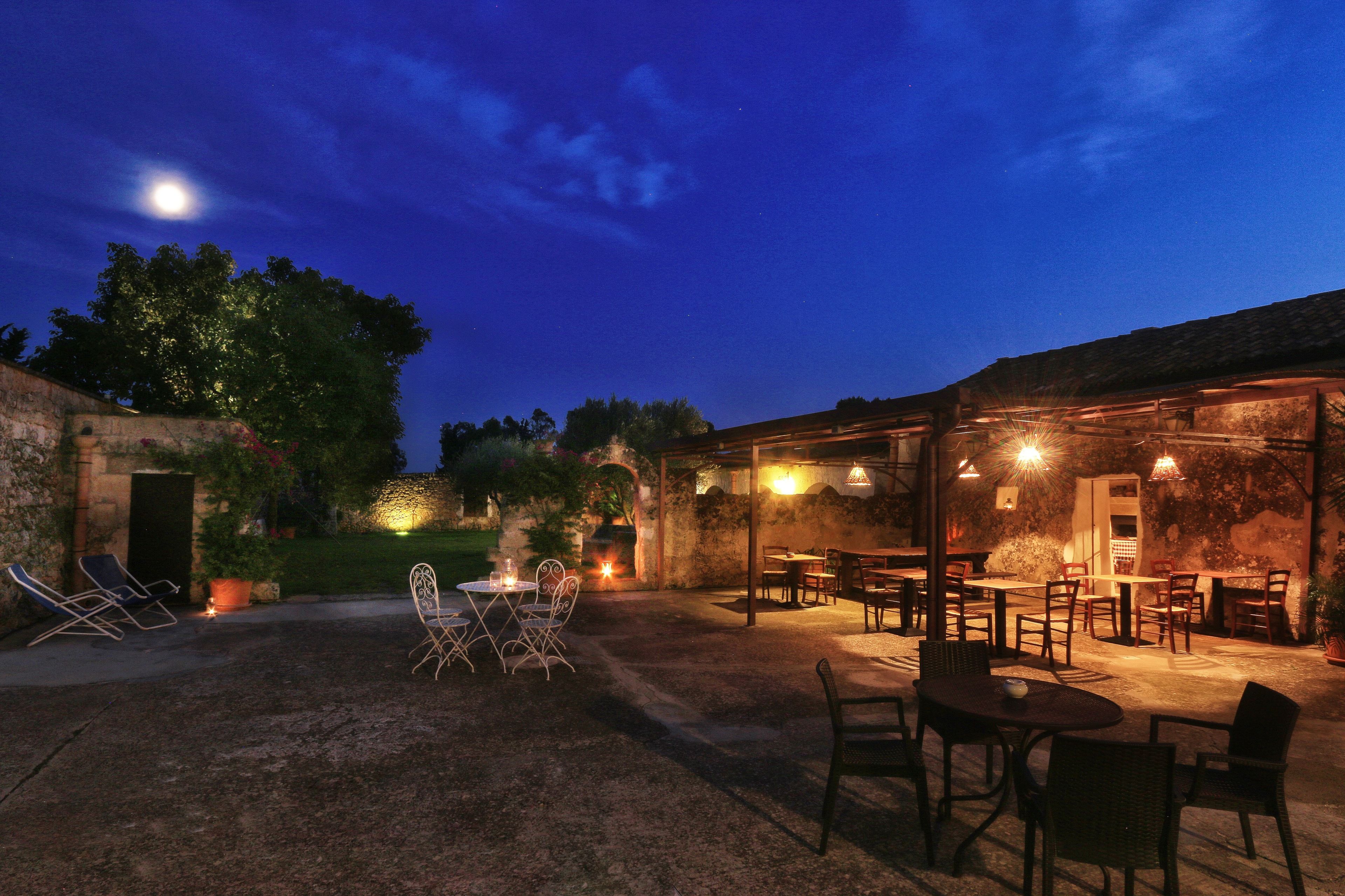Un'area patio all'aperto con tavoli e sedie, un muro in pietra e un pergolato con luci pendenti.