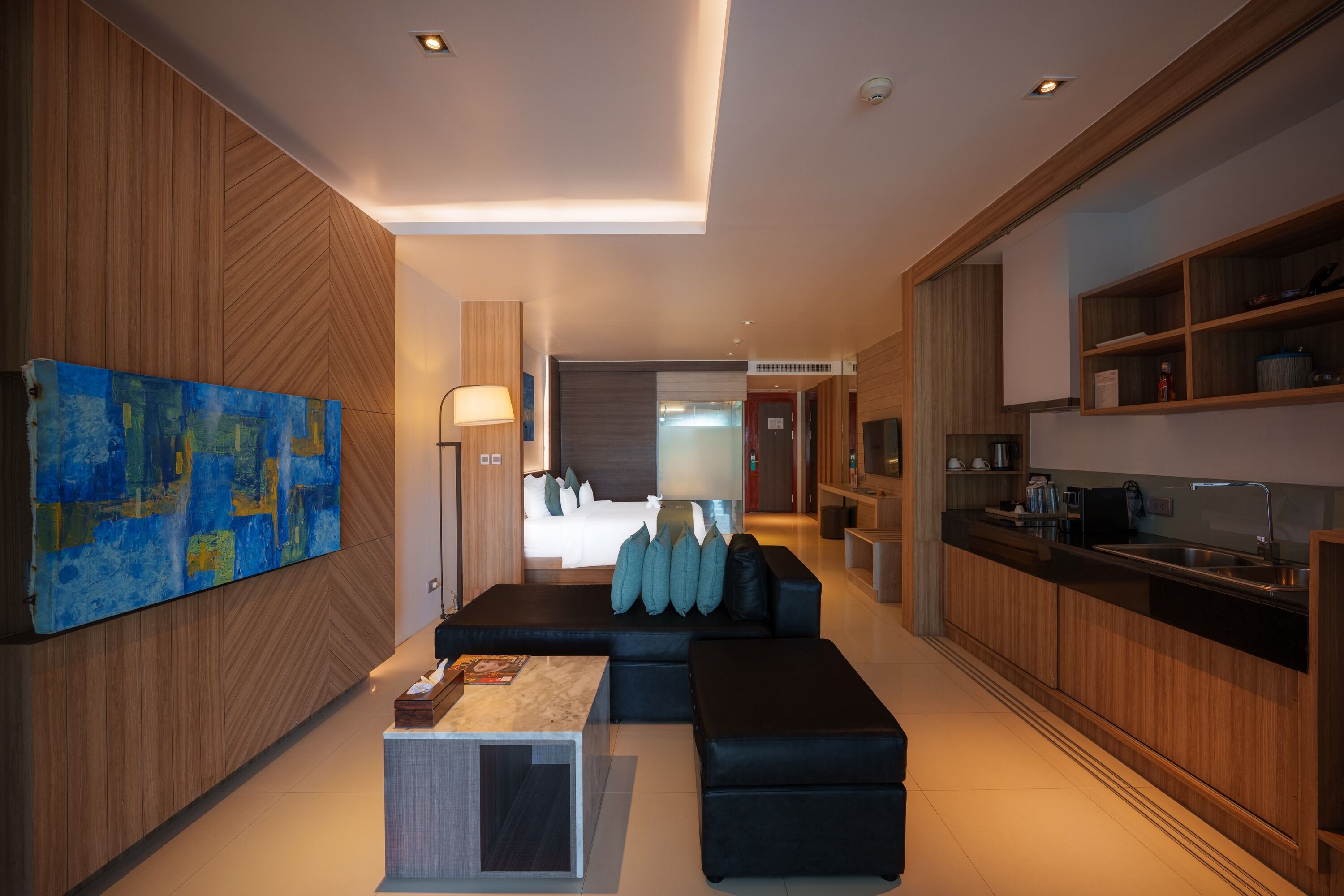 A modern hotel room with a large bed, a seating area with a coffee table, and a kitchen area with a sink and cabinets.
