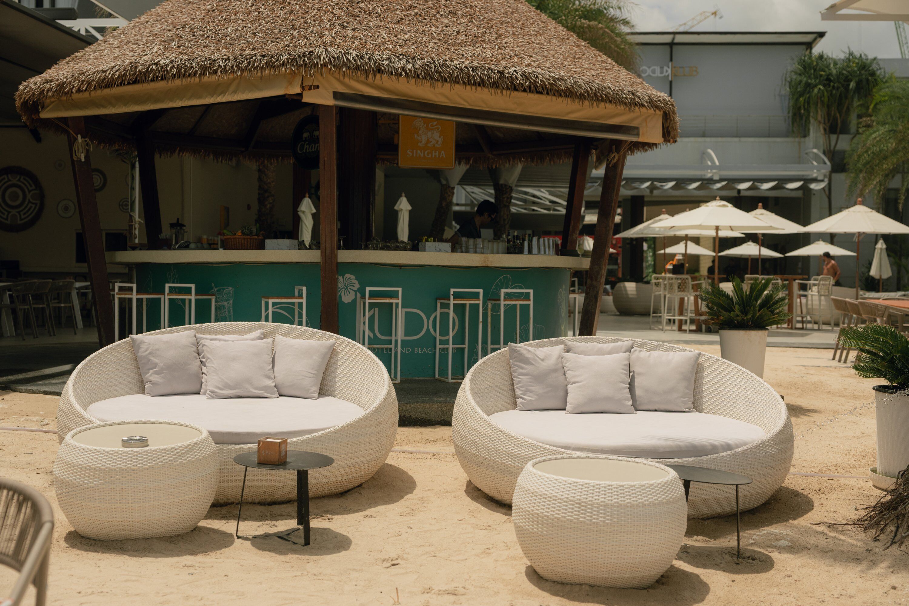 Outdoor wicker furniture with cushions, a thatched umbrella, and a bar area with a sign.
