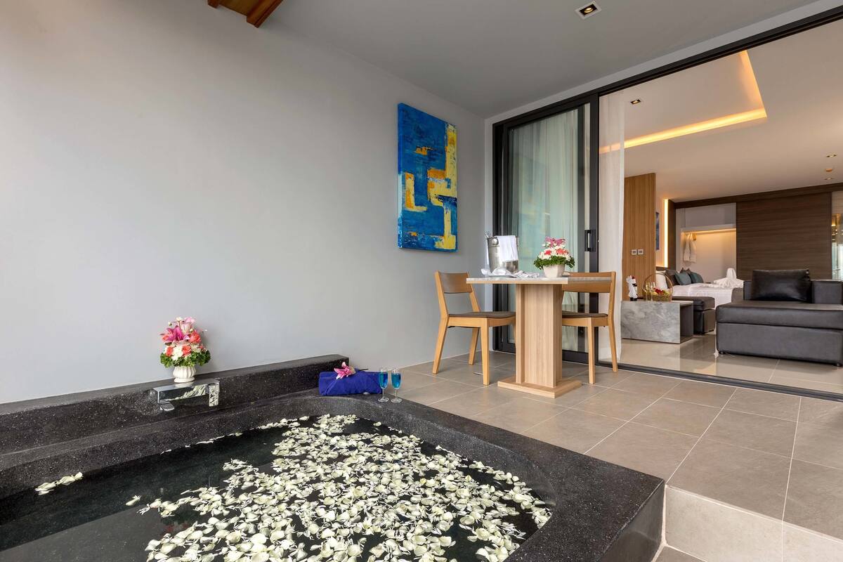 A modern living room with a black marble bathtub filled with white flowers, a wooden dining table with chairs, a blue abstract painting on the wall, and a grey sofa in the background.