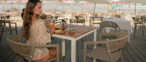Una persona sentada en una mesa con vista a la playa y sombrillas al fondo.