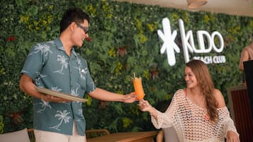 A person holding a drink with a straw and a plate, standing in front of a wall with greenery and a sign that reads 'KUDO BEACH CLUB'.