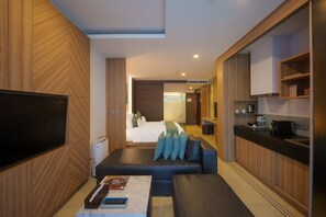 A modern hotel room with a large flat-screen TV, a sofa with cushions, a coffee table with books, and a kitchenette area. - Kudo Hotel & Beach Club (Adults Only) (Patong)