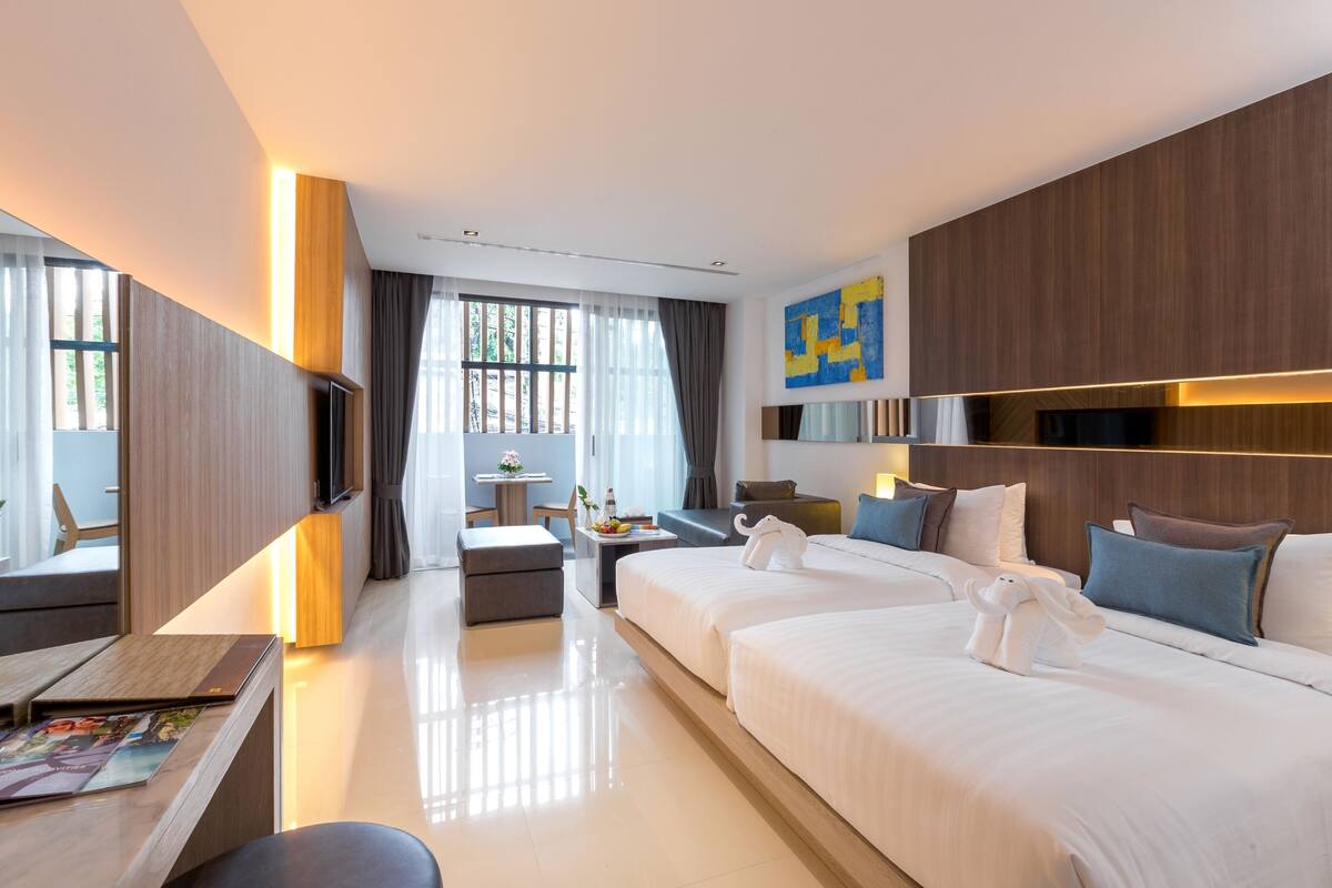 A modern hotel room with two beds, a desk, a chair, and a small table with a vase.