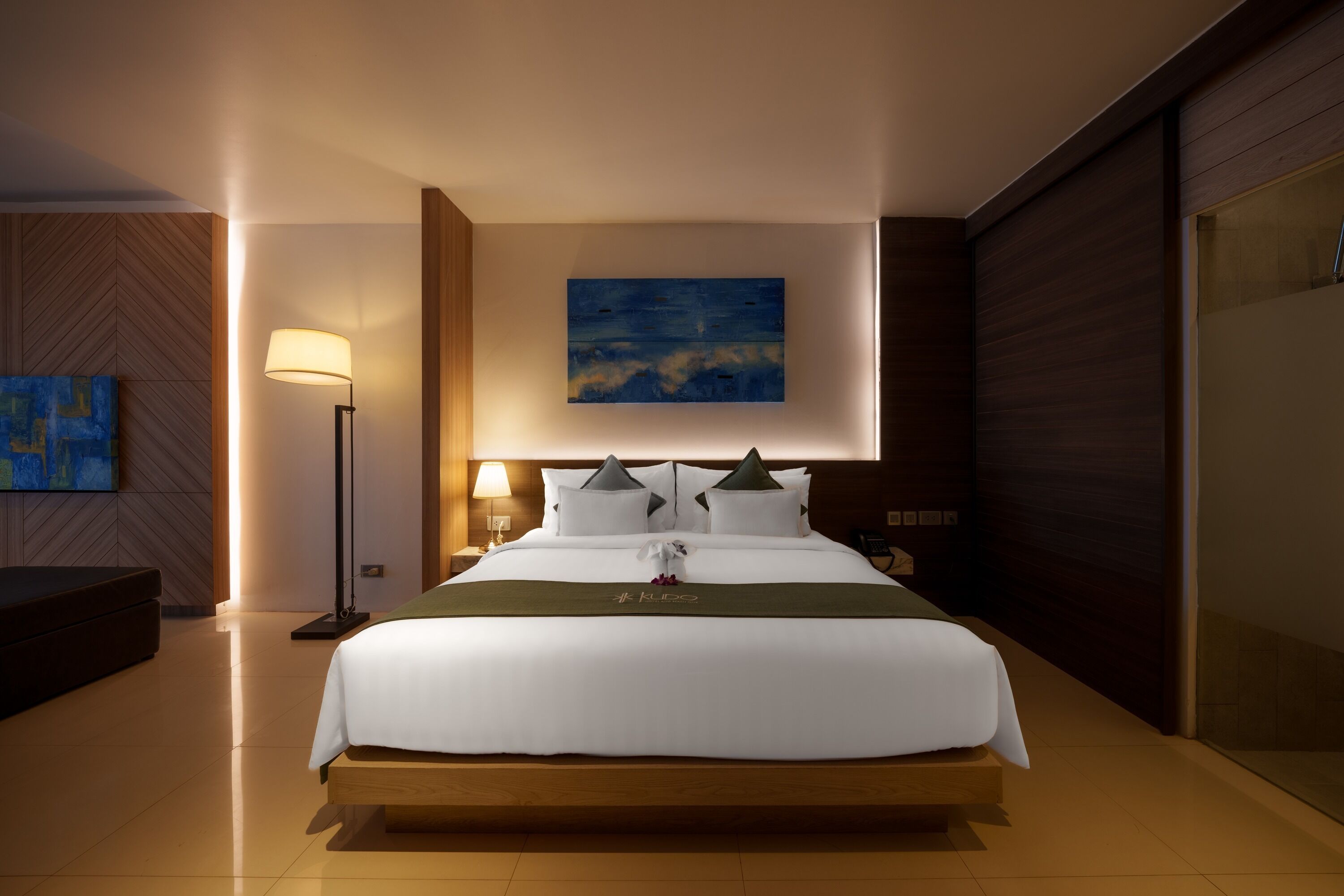 Ein Hotelzimmer mit einem großen Bett, einem Nachttisch mit Lampe, einem Gemälde an der Wand und Holzboden.