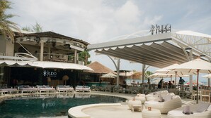 Un área de piscina con sillas de descanso y sombrillas, una zona de bar con asientos, y un letrero que dice 'KUDO BEACH CLUB'.