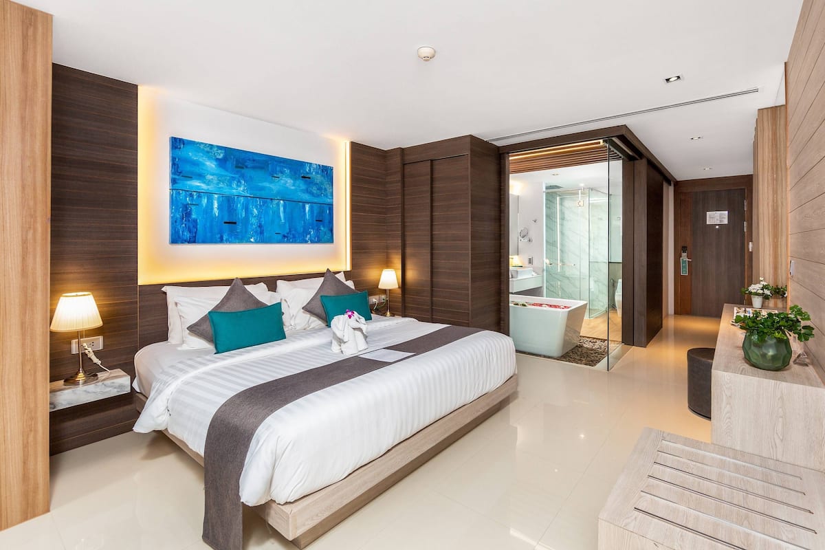 A modern hotel room with a large bed, two bedside tables with lamps, a large abstract painting, a glass-enclosed bathroom, and a wooden reception desk.