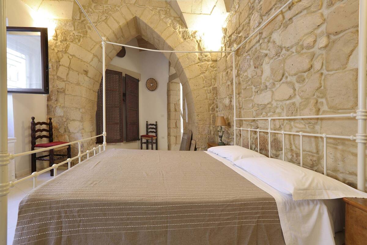 A room with a stone wall, a bed with a striped bedspread, a wooden chair, and a small table with a lamp.