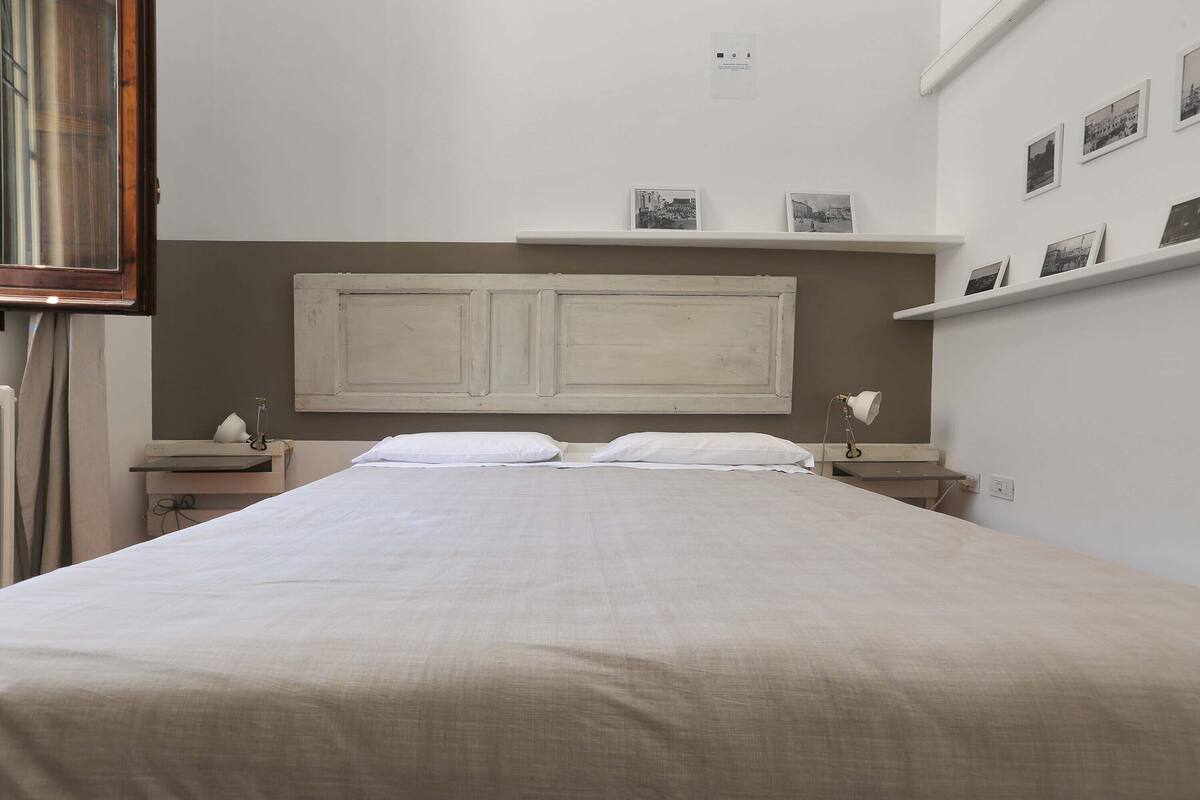 A bedroom with a large bed, two bedside tables, a nightstand, a wall-mounted shelf with pictures, and a window with curtains.
