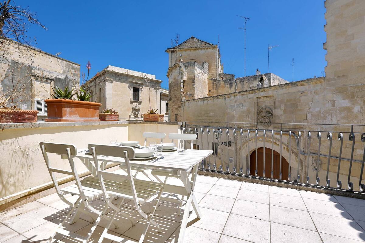 A terrace with a table and chairs, a potted plant, and a view of old buildings and a tower.