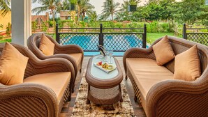A patio with wicker furniture, a dining table with a plate of food and glasses, and a view of a pool and greenery.