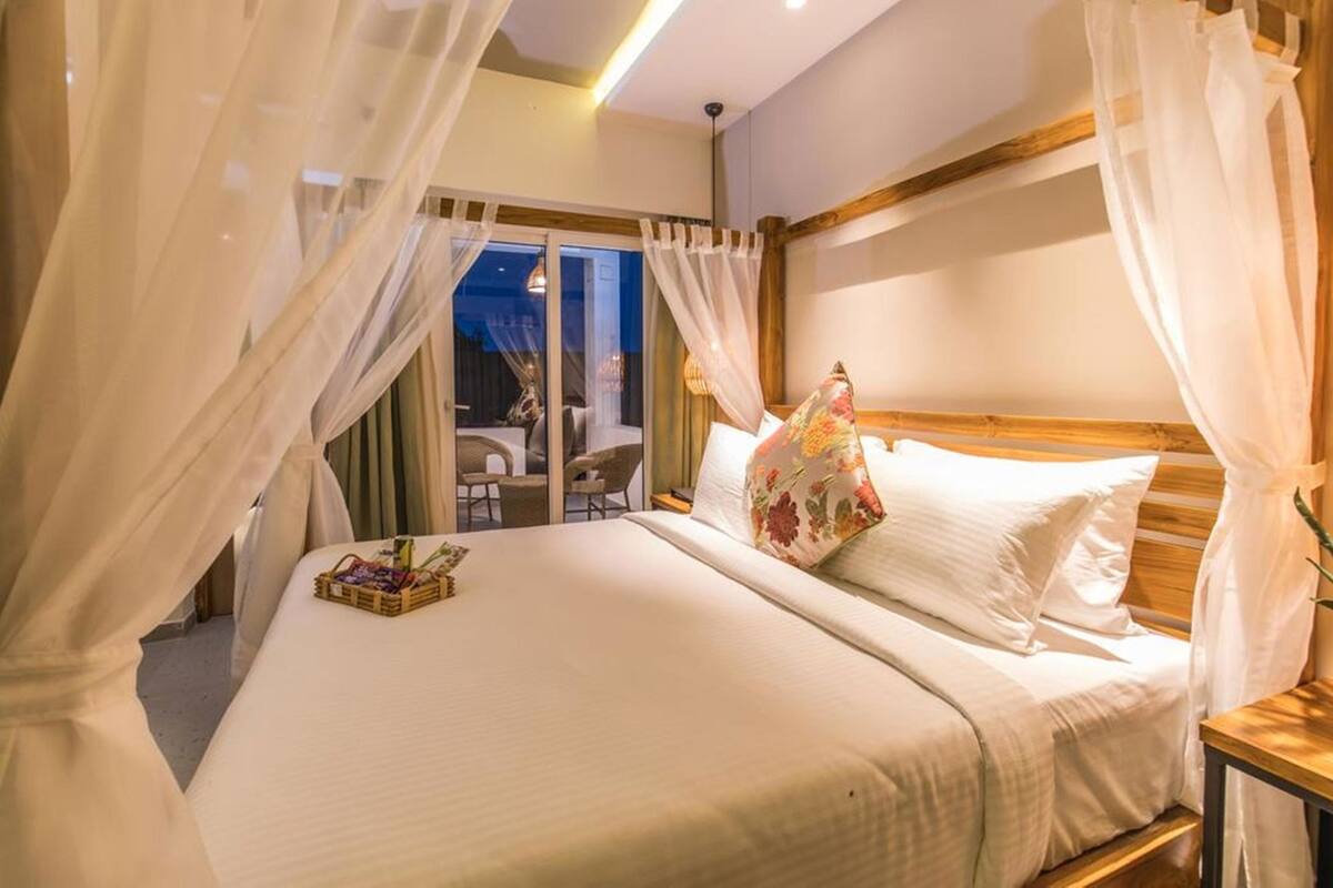 Habitación de hotel con cama, cortinas blancas, cabecera de madera y vistas al exterior.