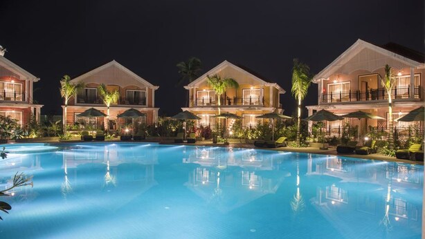 A swimming pool surrounded by villas with balconies, palm trees, and outdoor seating areas.