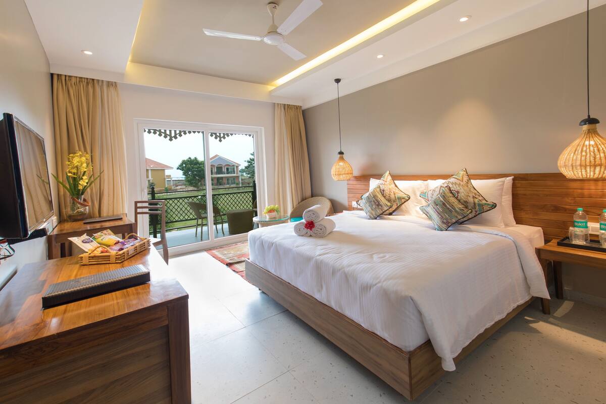Habitación de hotel con cama, escritorio, silla, televisión, ventana con cortinas y balcón.