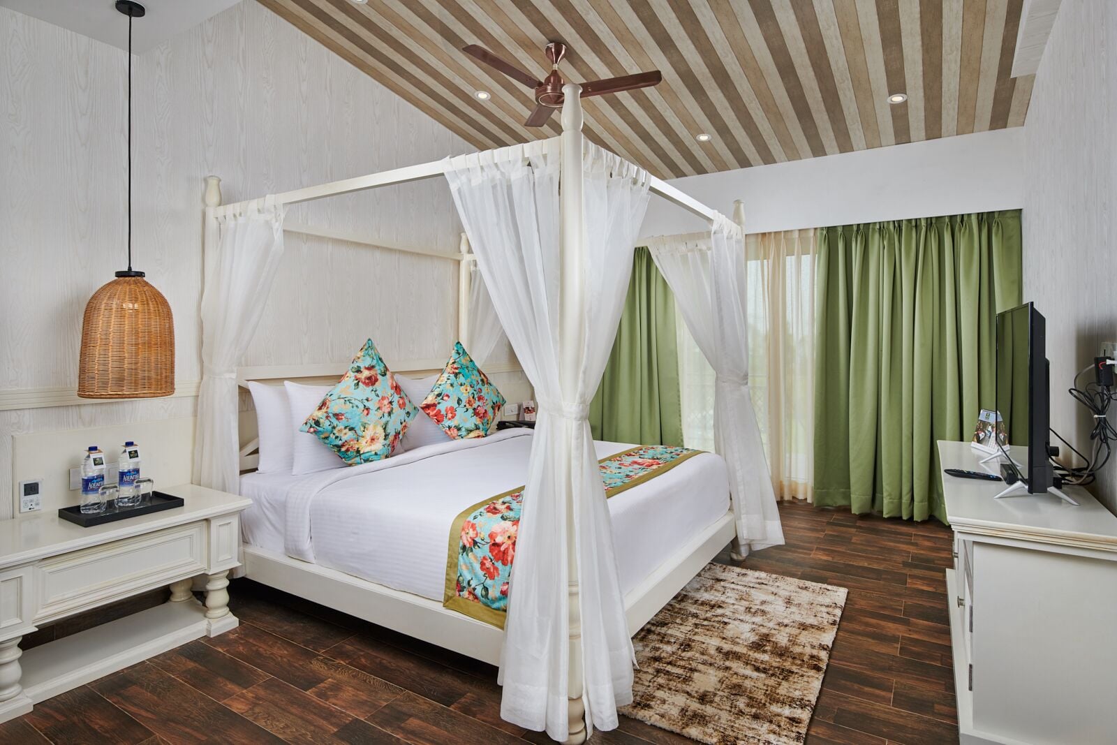 Quarto de hotel com cama de dossel, mesa de cabeceira com garrafa e copos, televisão em suporte e chão de madeira.
