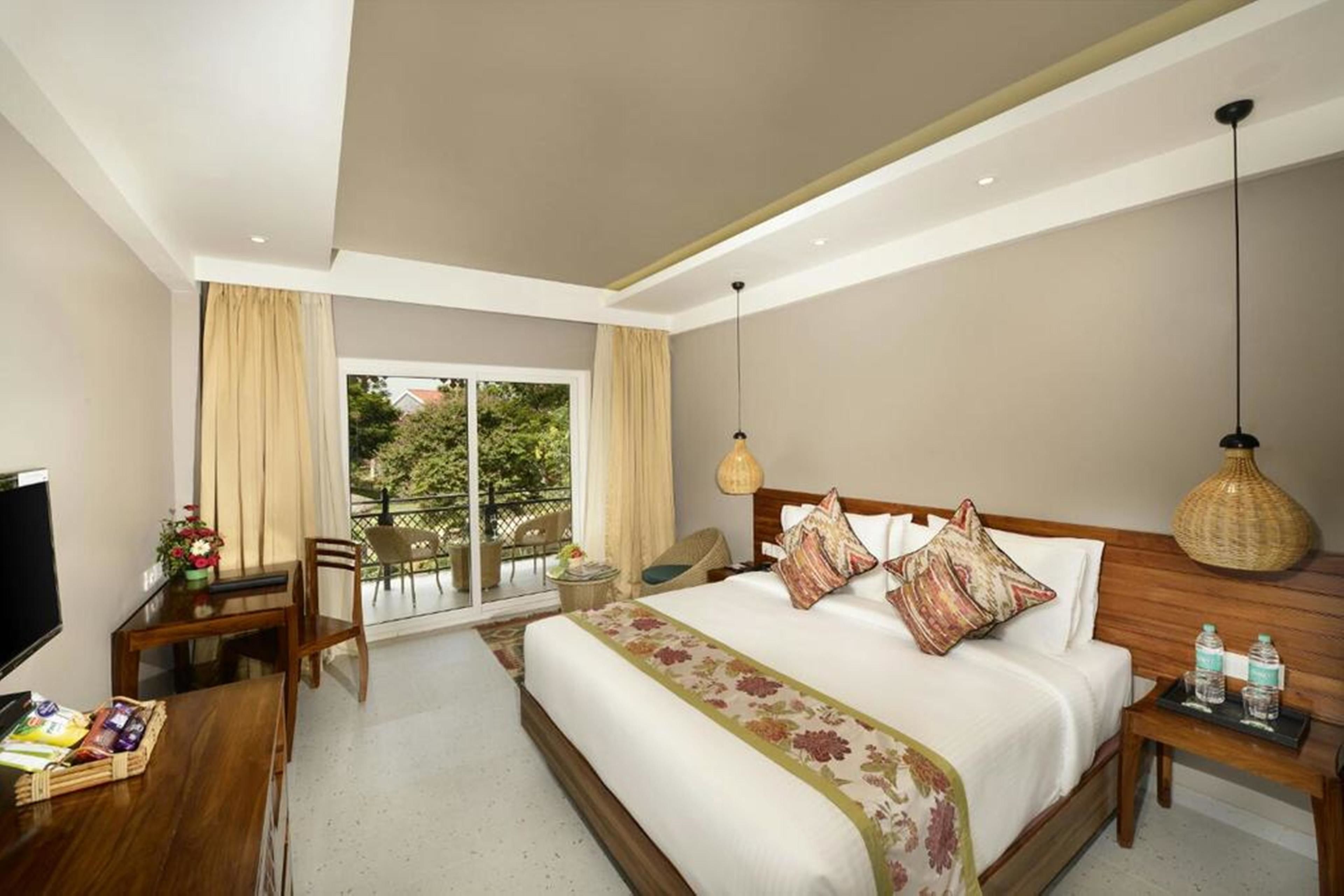 A hotel room with a large bed, a TV, a desk with a chair, and a balcony with outdoor seating.