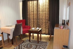 Quarto de hotel com uma mesa de jantar, duas cadeiras, uma pequena escrivaninha de madeira e uma janela grande com cortinas.
