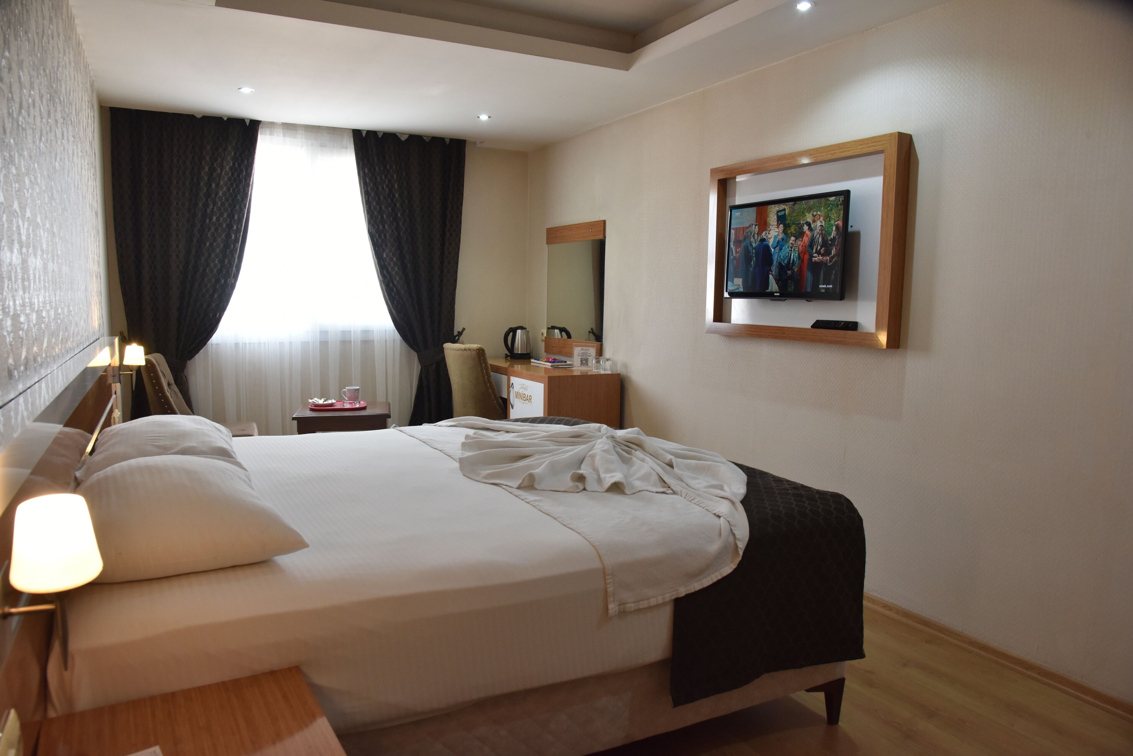 Habitación de hotel con cama, escritorio con lámpara, silla, cafetera, un cuadro en la pared y ventana con cortinas.