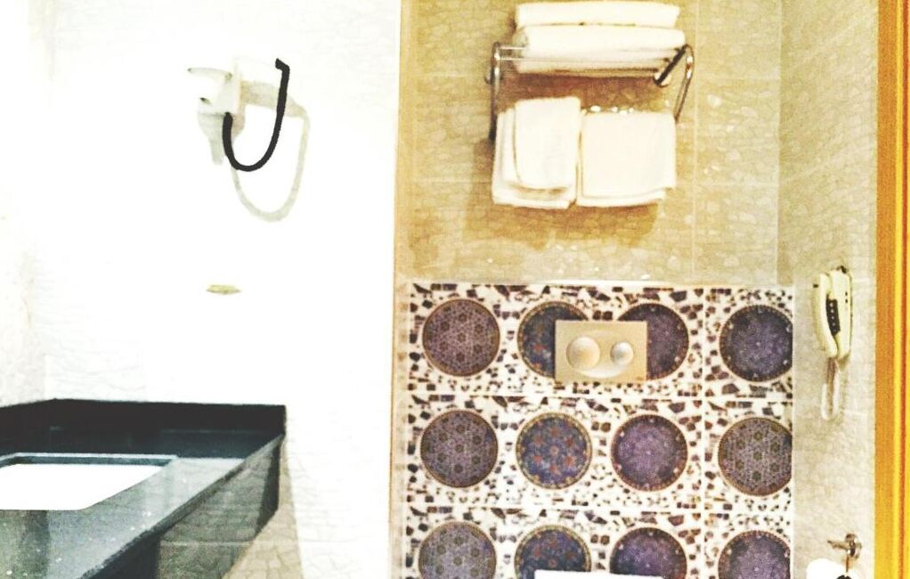 Un baño con lavamanos, un perchero con toallas, un inodoro con un salpicadero de azulejos estampados y un teléfono.