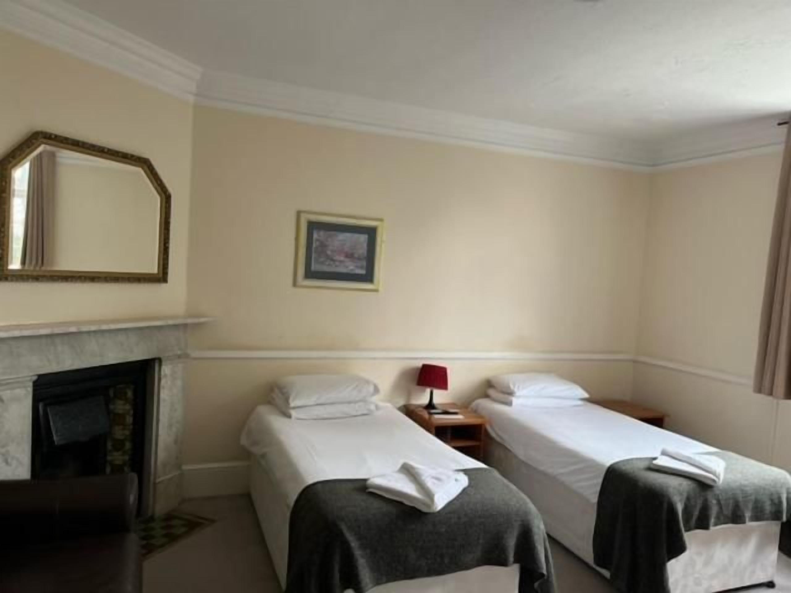 A hotel room with two single beds, a small table, a chair, a nightstand with a lamp, and a fireplace.
