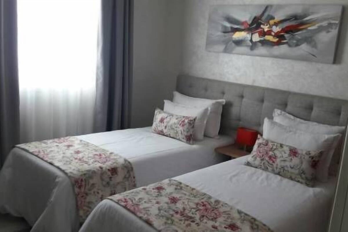 A hotel room with two beds, each with floral bedspreads and white pillows. There is a grey headboard, a painting on the wall, and a window with curtains.