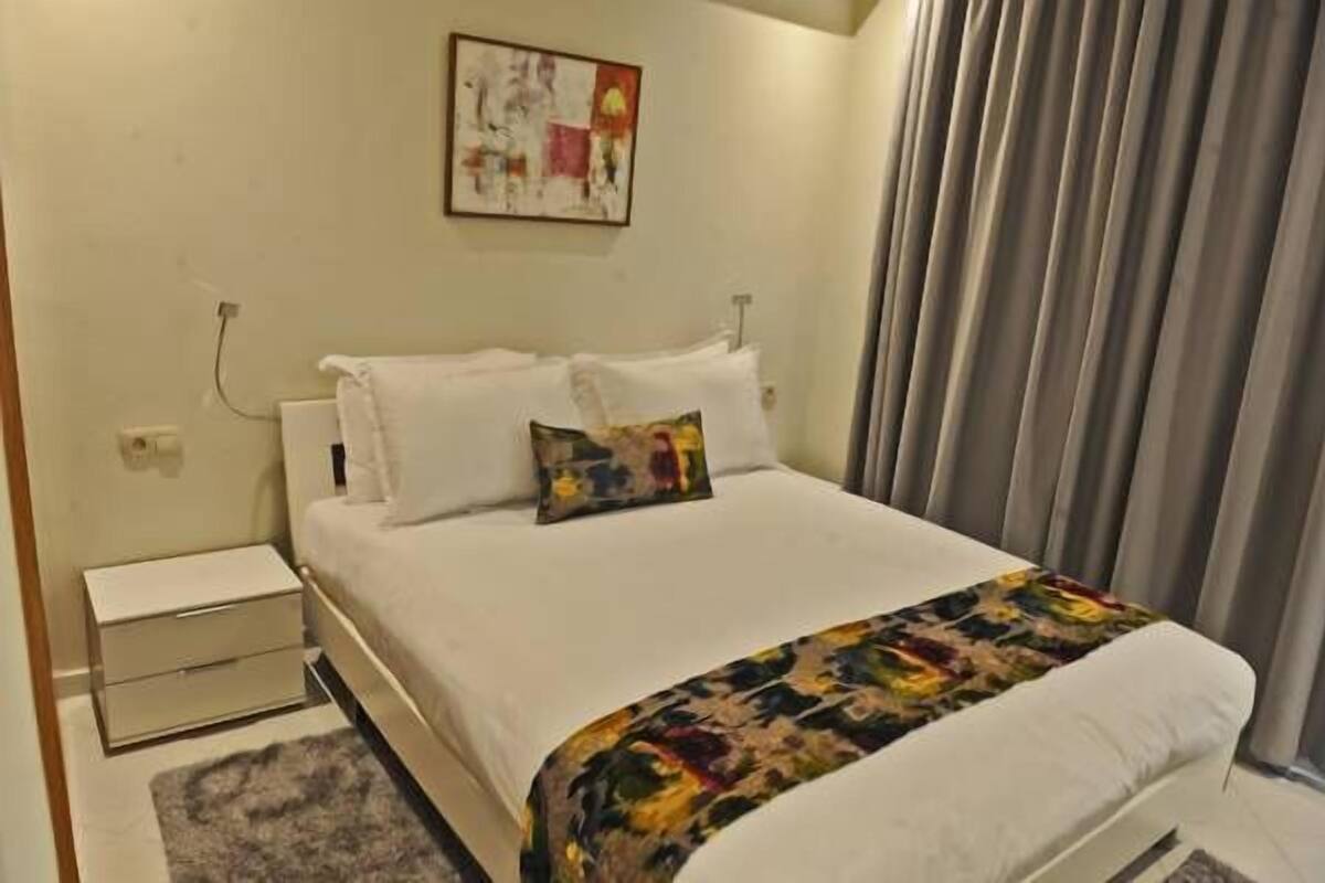A neatly made bed with white linens and a colorful throw blanket, a small bedside table, and a painting on the wall.