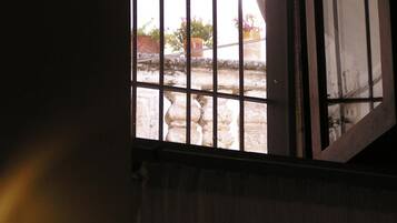 Ein Fenster mit Metallgitter, einem halb heruntergezogenen Vorhang und Blick auf Pflanzen und eine Steinmauer im Freien.