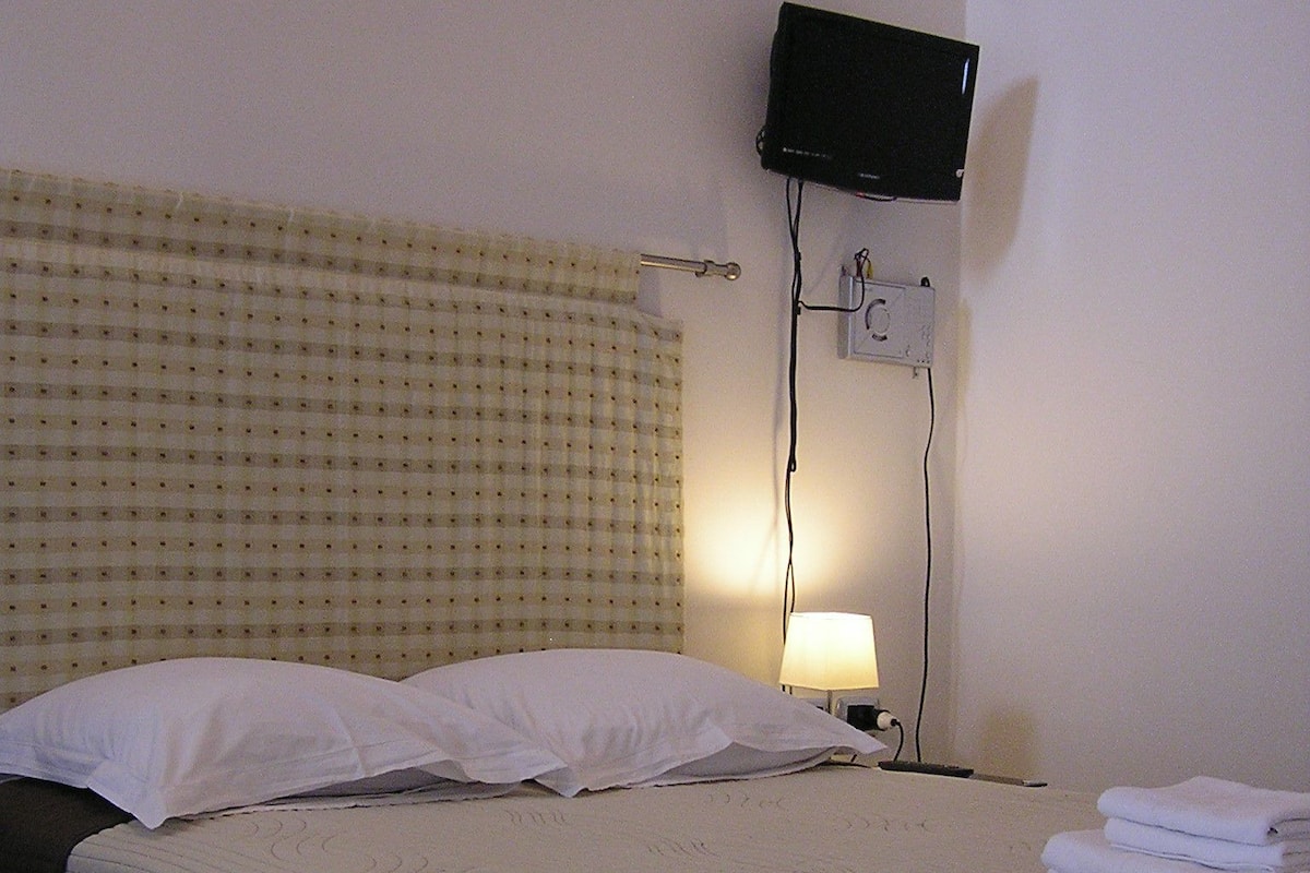 A hotel room with a bed, pillows, a TV mounted on the wall, and a bedside table with a lamp.