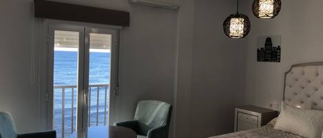 Habitación de hotel con cama, mesa, dos sillas, mesita de noche y vista al mar a través de una puerta corrediza.