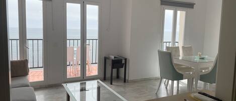 A modern living room with a glass coffee table, a dining area with a glass table, and a balcony with a view of the sea.
